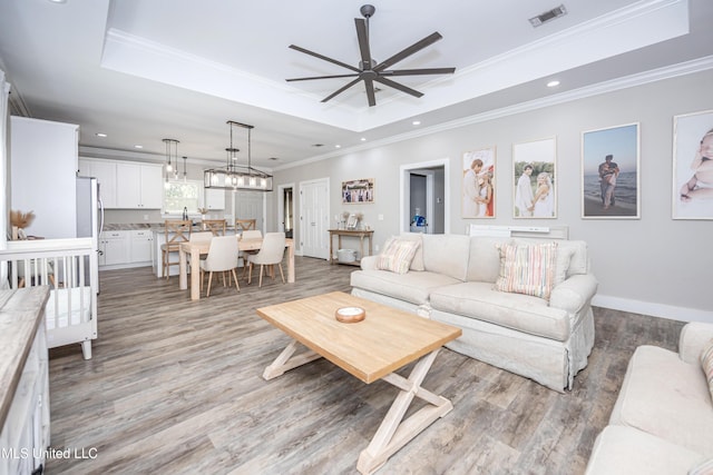 living room with ceiling fan with notable chandelier, ornamental molding, a raised ceiling, and light wood-type flooring