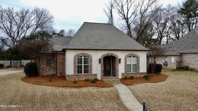 french country home with a shingled roof, fence, and brick siding