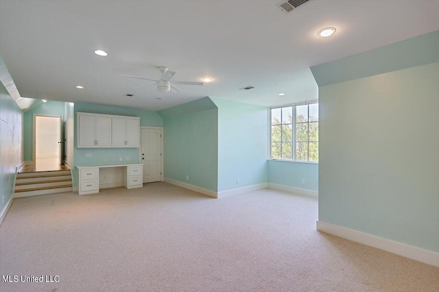 unfurnished living room with built in desk, light colored carpet, and ceiling fan