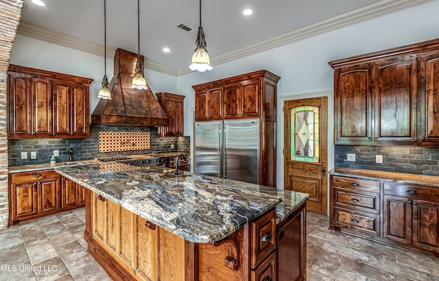 kitchen with stainless steel appliances, dark stone counters, an island with sink, and custom exhaust hood