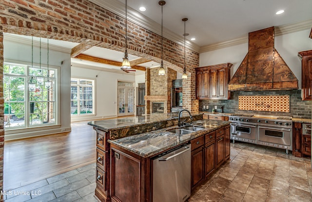 kitchen with sink, stainless steel appliances, decorative light fixtures, premium range hood, and a kitchen island with sink