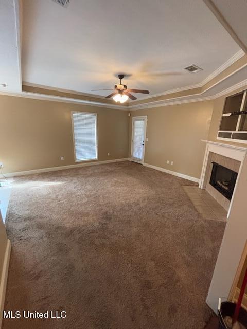 unfurnished living room featuring carpet floors, ceiling fan, a tray ceiling, a tile fireplace, and crown molding