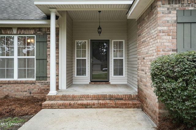 view of exterior entry featuring covered porch, brick siding, and roof with shingles