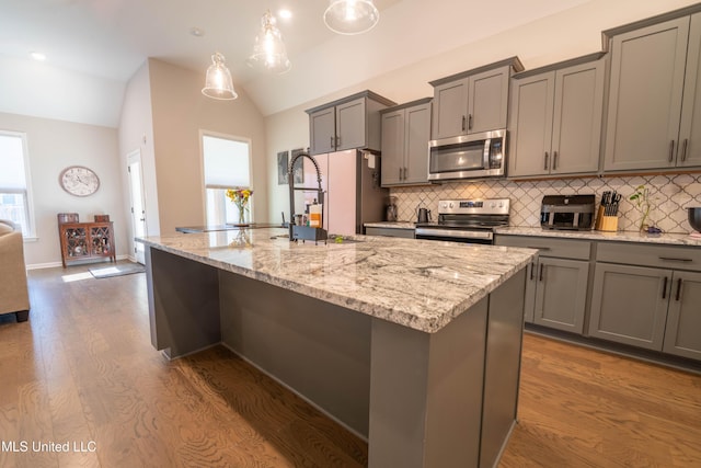 kitchen featuring decorative light fixtures, vaulted ceiling, an island with sink, wood-type flooring, and appliances with stainless steel finishes