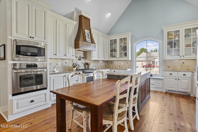 kitchen featuring light wood-style flooring, glass insert cabinets, light stone counters, custom exhaust hood, and stainless steel appliances