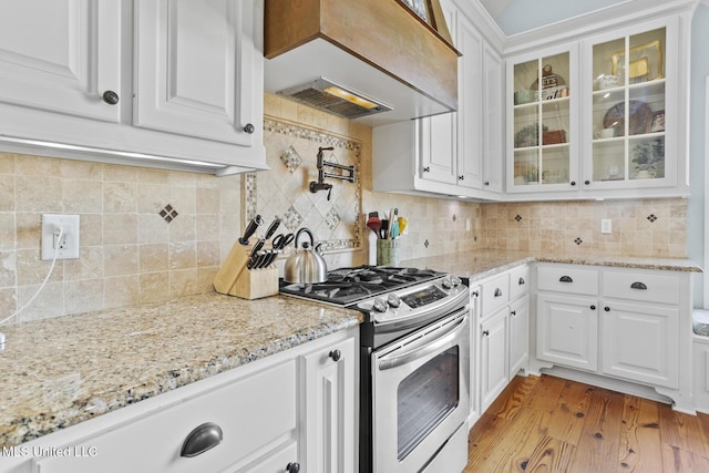 kitchen with premium range hood, stainless steel range with gas stovetop, white cabinetry, light wood-style floors, and glass insert cabinets