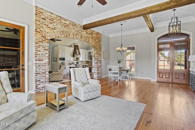 living room featuring ornamental molding, beamed ceiling, french doors, light wood-style floors, and ceiling fan with notable chandelier