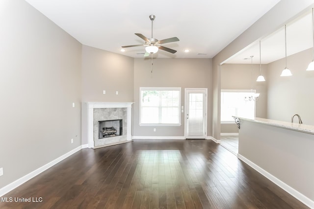 unfurnished living room featuring a ceiling fan, a chandelier, a tiled fireplace, baseboards, and dark wood finished floors