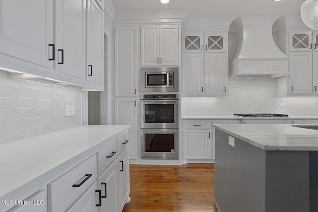 kitchen with stainless steel appliances, premium range hood, wood finished floors, white cabinets, and backsplash