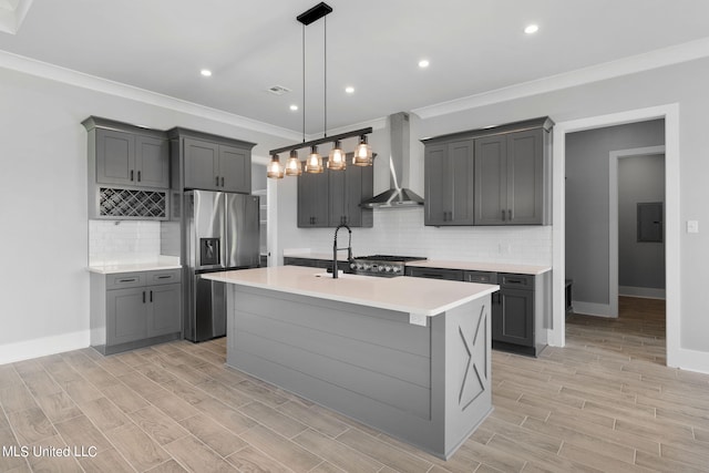 kitchen with wall chimney range hood, appliances with stainless steel finishes, a kitchen island with sink, sink, and decorative light fixtures