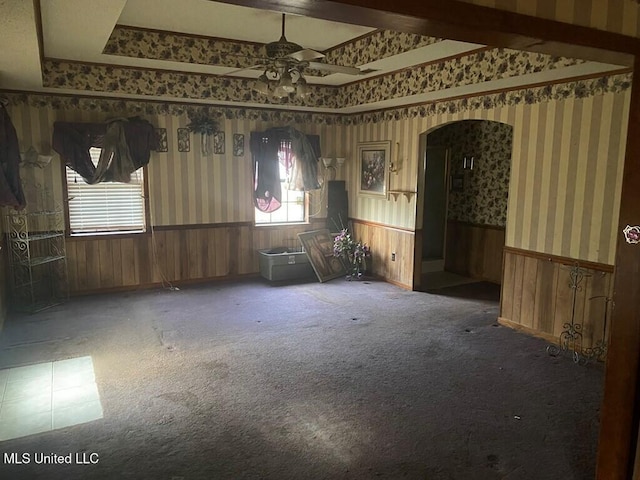 carpeted empty room featuring wallpapered walls, arched walkways, a ceiling fan, a wainscoted wall, and a tray ceiling