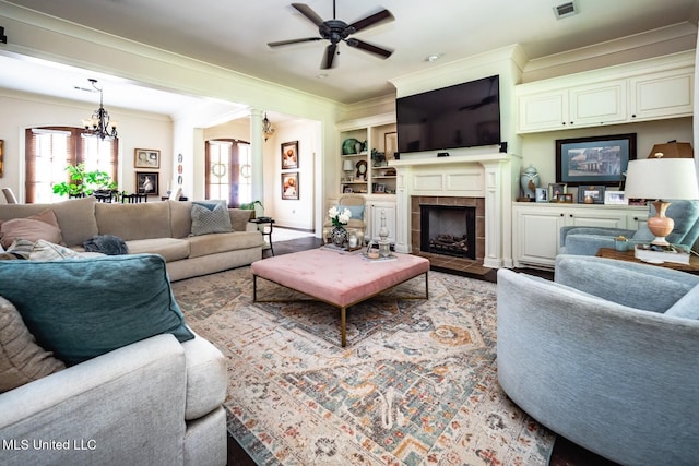 living room with a tiled fireplace, ornamental molding, ceiling fan with notable chandelier, and built in features