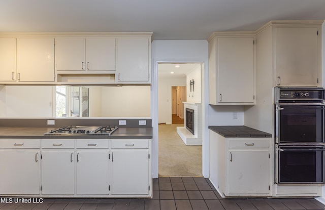 kitchen with tile countertops, dobule oven black, a brick fireplace, dark colored carpet, and stainless steel gas stovetop