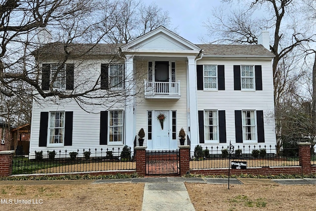 greek revival house featuring a balcony, a fenced front yard, a chimney, and a shingled roof