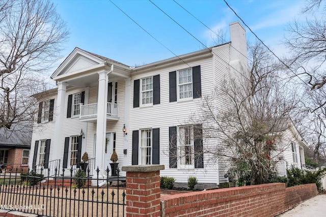 neoclassical home featuring a balcony, a fenced front yard, and a chimney