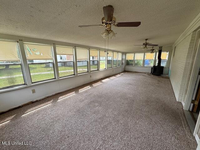 unfurnished sunroom featuring a wood stove, carpet, ceiling fan, and a textured ceiling