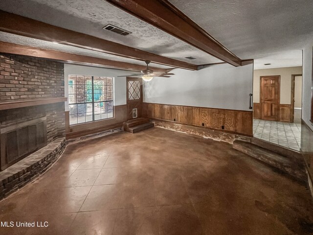 unfurnished living room featuring wood walls, beam ceiling, a brick fireplace, a textured ceiling, and ceiling fan