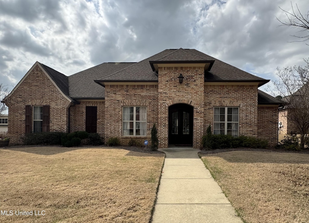 french country inspired facade featuring brick siding, a front yard, and roof with shingles