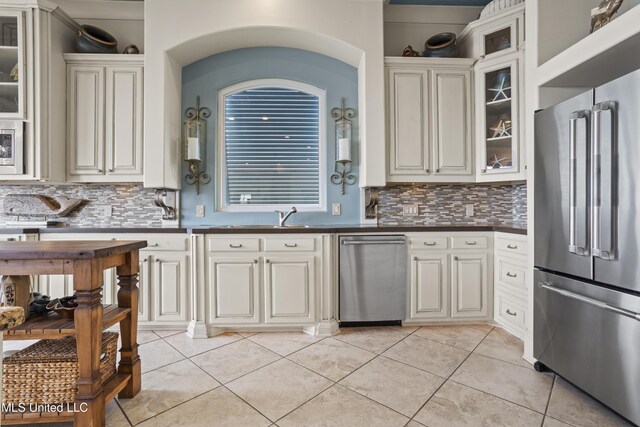 kitchen featuring stainless steel appliances, backsplash, dark countertops, light tile patterned floors, and glass insert cabinets