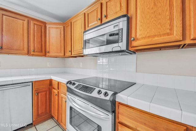 kitchen with appliances with stainless steel finishes, tile counters, ornamental molding, and light tile patterned flooring