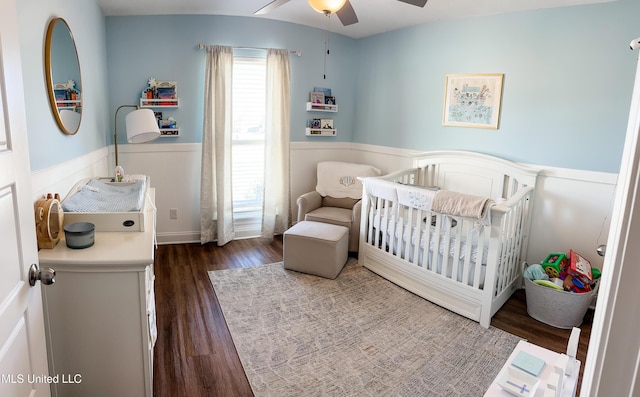 bedroom featuring dark wood-type flooring, wainscoting, ceiling fan, and a crib