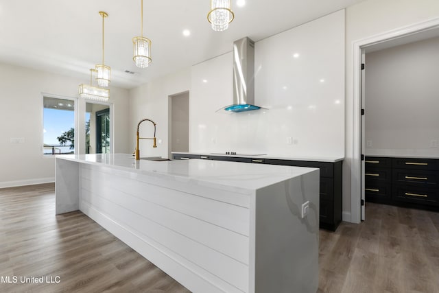 kitchen featuring an inviting chandelier, hanging light fixtures, wall chimney exhaust hood, an island with sink, and wood-type flooring