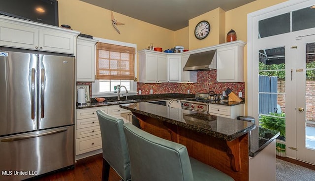 kitchen with a kitchen breakfast bar, dark stone counters, wall chimney exhaust hood, stainless steel appliances, and white cabinets