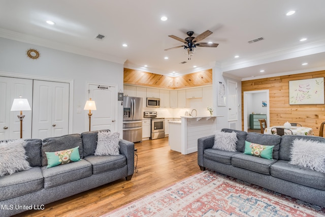 living room with ceiling fan, light wood-type flooring, sink, crown molding, and wooden walls