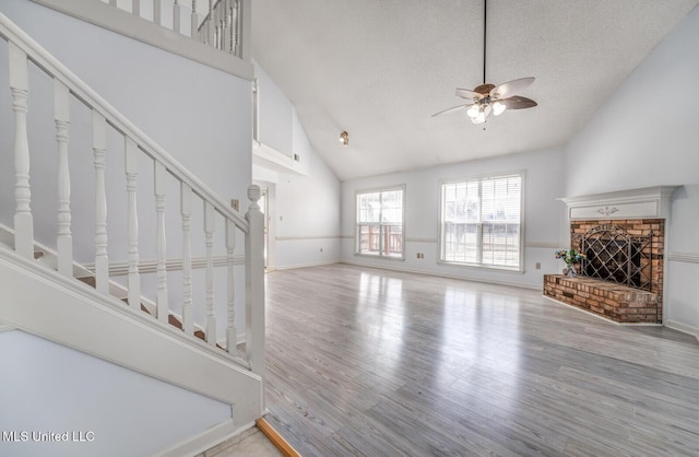 unfurnished living room with high vaulted ceiling, a brick fireplace, ceiling fan, light wood-type flooring, and a textured ceiling