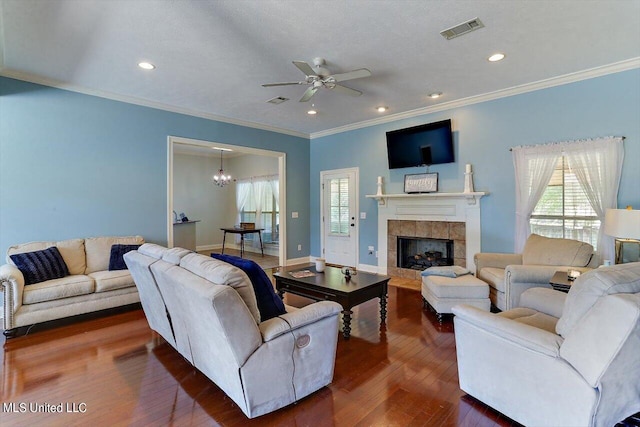 living room featuring crown molding, dark hardwood / wood-style floors, a tiled fireplace, and ceiling fan with notable chandelier