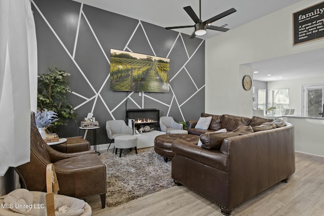living room featuring a lit fireplace, wood finished floors, a ceiling fan, and an accent wall