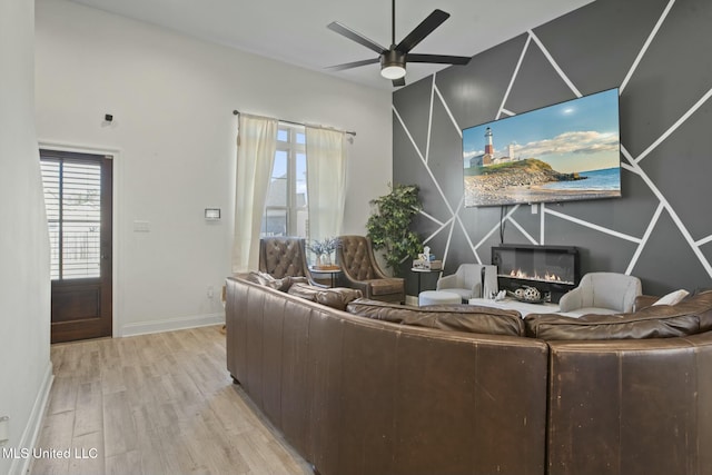 living area featuring a ceiling fan, a glass covered fireplace, baseboards, and light wood finished floors