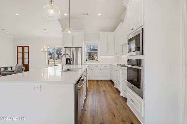 kitchen with an island with sink, appliances with stainless steel finishes, light countertops, white cabinetry, and pendant lighting