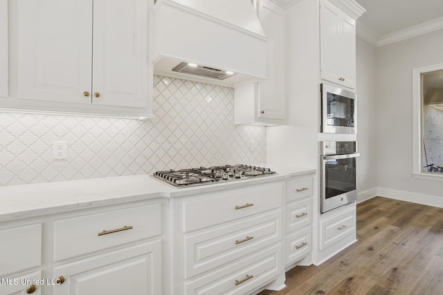 kitchen featuring light stone counters, custom exhaust hood, stainless steel appliances, ornamental molding, and white cabinets