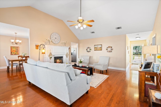 living area with visible vents, high vaulted ceiling, ceiling fan with notable chandelier, a warm lit fireplace, and wood finished floors