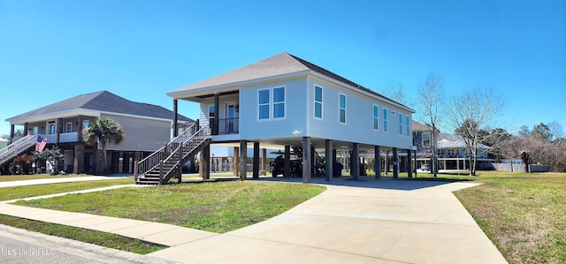 raised beach house featuring covered porch, concrete driveway, stairway, a carport, and a front lawn