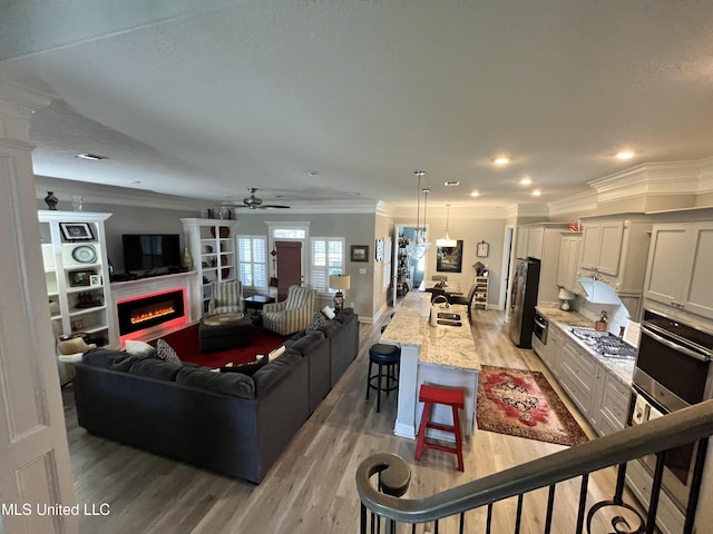 living room with light wood-style floors, a glass covered fireplace, a ceiling fan, baseboards, and ornamental molding