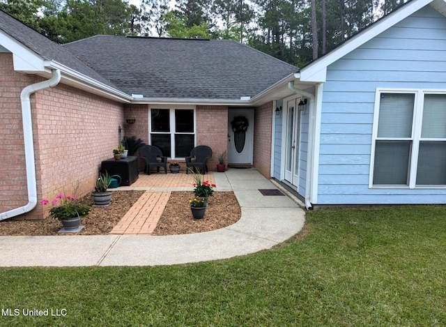 property entrance with brick siding, a shingled roof, a patio area, and a yard
