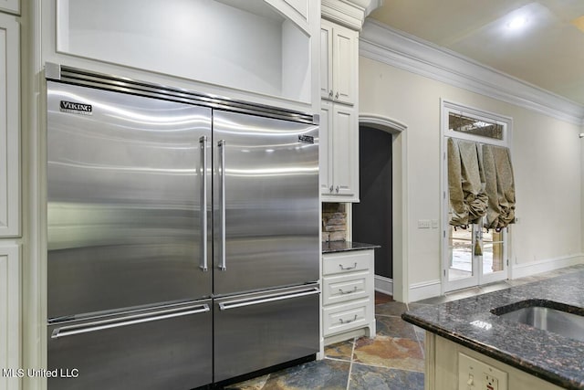 kitchen with white cabinets, dark stone counters, built in refrigerator, crown molding, and a healthy amount of sunlight