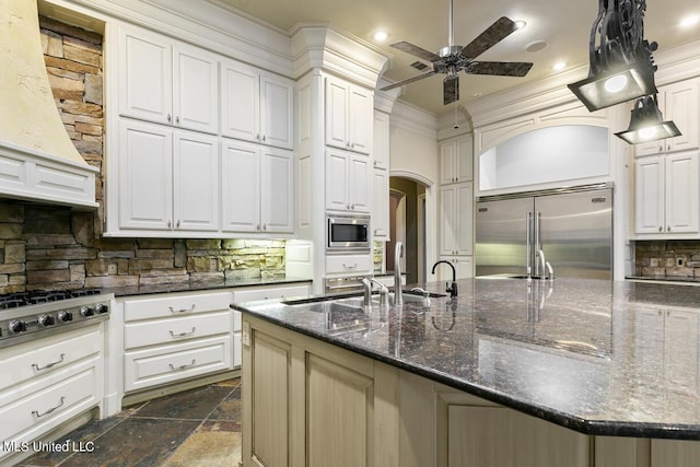 kitchen with built in appliances, a center island with sink, dark stone countertops, decorative backsplash, and white cabinets