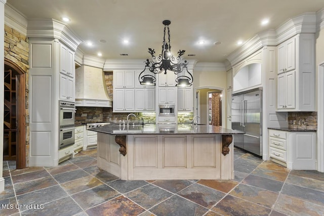 kitchen featuring premium range hood, built in appliances, a center island with sink, a kitchen breakfast bar, and white cabinets