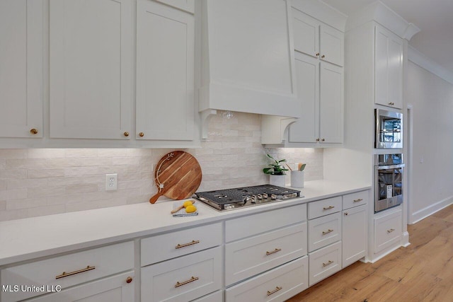 kitchen with appliances with stainless steel finishes, light hardwood / wood-style flooring, tasteful backsplash, custom range hood, and white cabinets