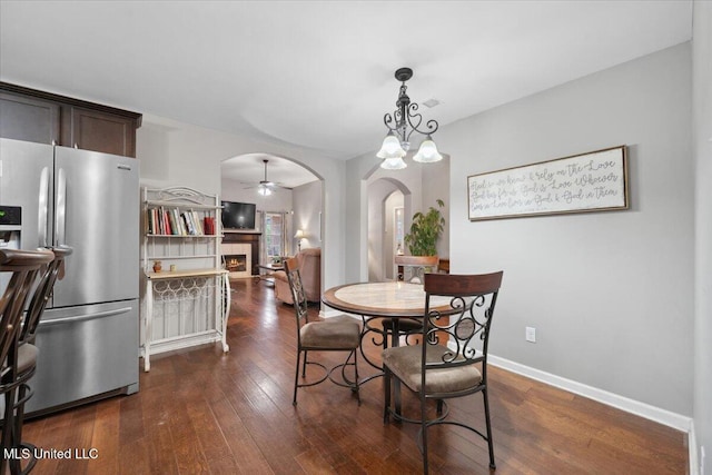 dining area with arched walkways, a ceiling fan, a warm lit fireplace, dark wood-style flooring, and baseboards