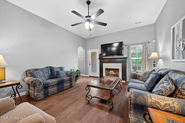 living room featuring arched walkways, a ceiling fan, wood finished floors, a tiled fireplace, and baseboards