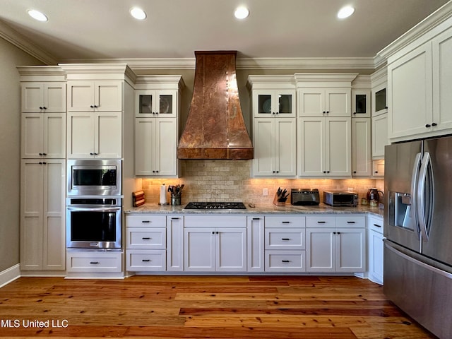 kitchen featuring stainless steel appliances, ornamental molding, premium range hood, light hardwood / wood-style floors, and tasteful backsplash