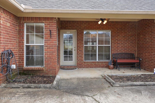 view of exterior entry with ceiling fan, a patio, brick siding, and roof with shingles