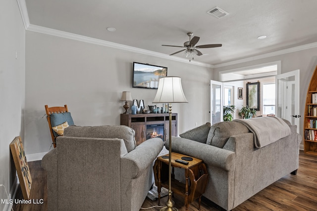 living area featuring ceiling fan, dark wood finished floors, visible vents, ornamental molding, and a glass covered fireplace