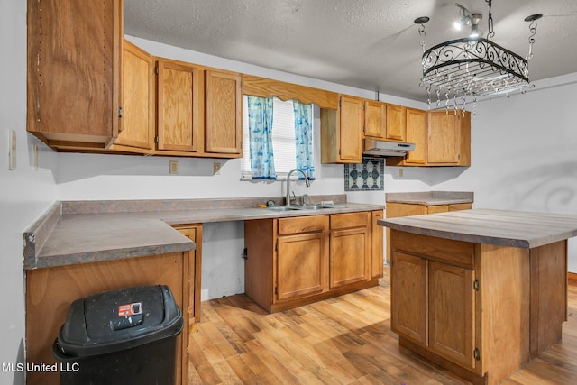 kitchen with a sink, under cabinet range hood, light wood finished floors, brown cabinetry, and a textured ceiling