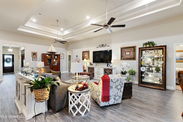 living area featuring ceiling fan, wood finished floors, baseboards, ornamental molding, and a tray ceiling