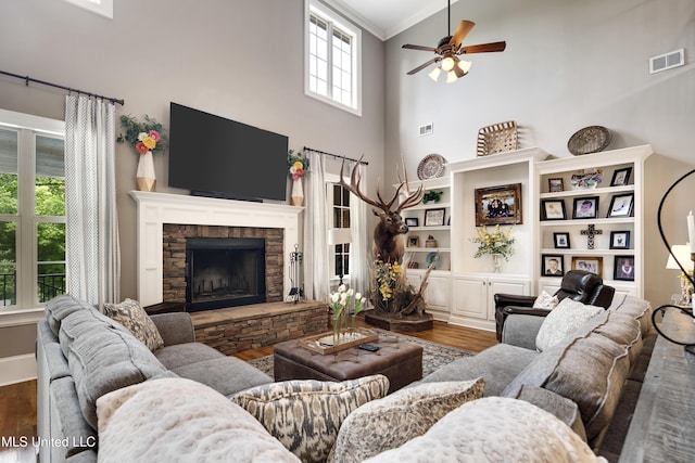 living area featuring wood finished floors, a ceiling fan, a high ceiling, a stone fireplace, and ornamental molding